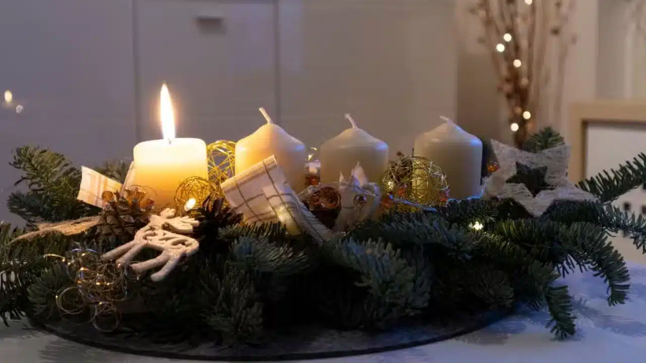 Adventný veniec s jednou zapálenou sviečkou, ozdobený zelenými vetvičkami, šiškami, zlatými ozdobami a dekoratívnymi prvkami, položený na stole v útulnom interiéri.
