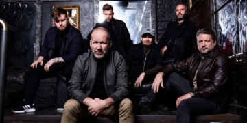 Skupina David Koller Band v industriálnom prostredí, členovia sedia a stoja v tmavom, modernom priestore.