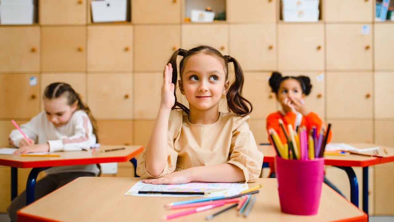 Dievčatko s dvomi copmi zdvihnutou rukou v triede, obklopené školskými pomôckami, zatiaľ čo ostatné deti pracujú.