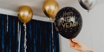Čierno-zlatý balón s nápisom "Happy New Year!" na Silvestrovskú oslavu, ozdobený bodkami, v interiéri s ďalšími zlatými a striebornými balónmi v pozadí.