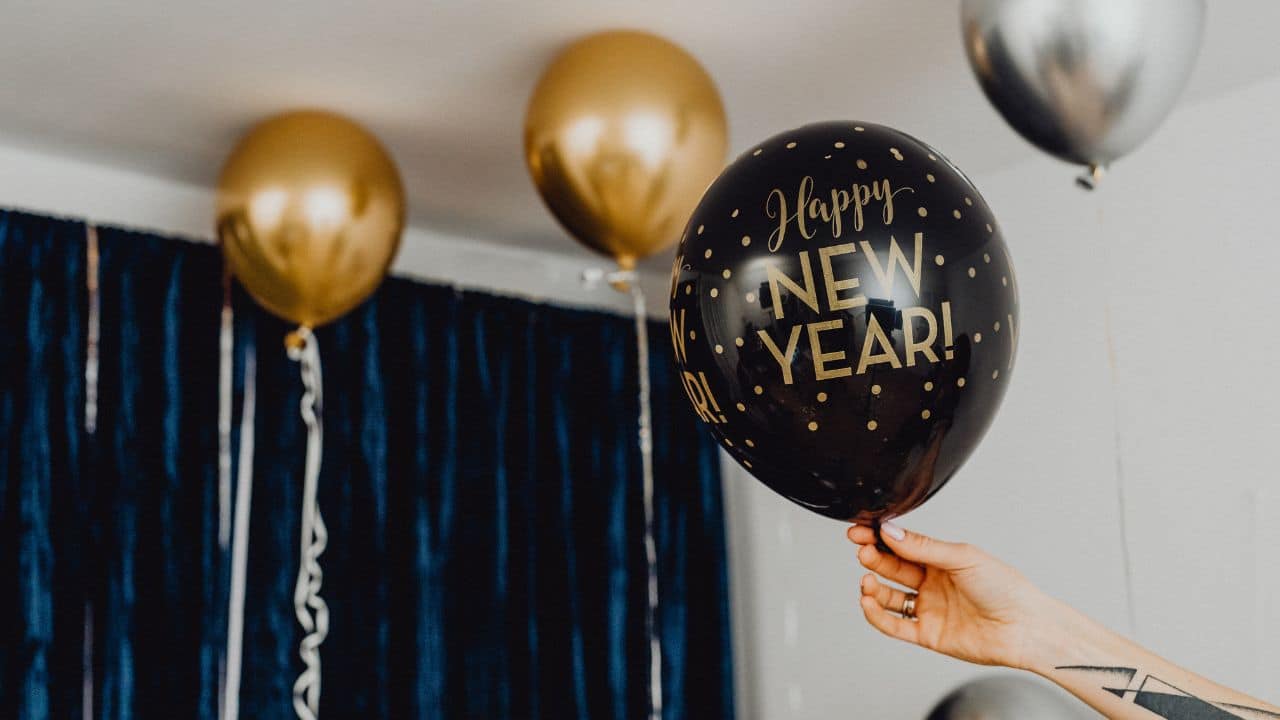 Čierno-zlatý balón s nápisom "Happy New Year!" na Silvestrovskú oslavu, ozdobený bodkami, v interiéri s ďalšími zlatými a striebornými balónmi v pozadí.