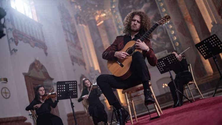 Gitarista Dávid Bílek počas vystúpenia s klasickou gitarou v historickom priestore, sprevádzaný sláčikovým kvartetom.