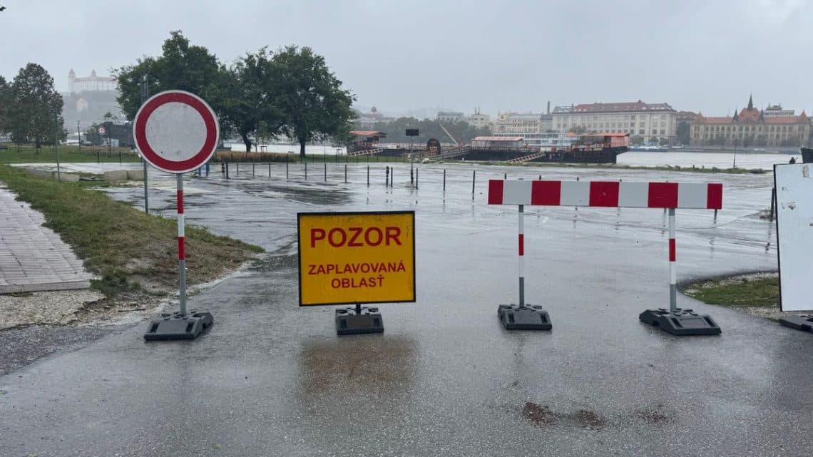Záplavová oblasť označená dopravnou značkou a varovným žltým nápisom 'Pozor: zaplavovaná oblasť,' s pohľadom na zaplavenú cestu a Bratislavský hrad v pozadí.