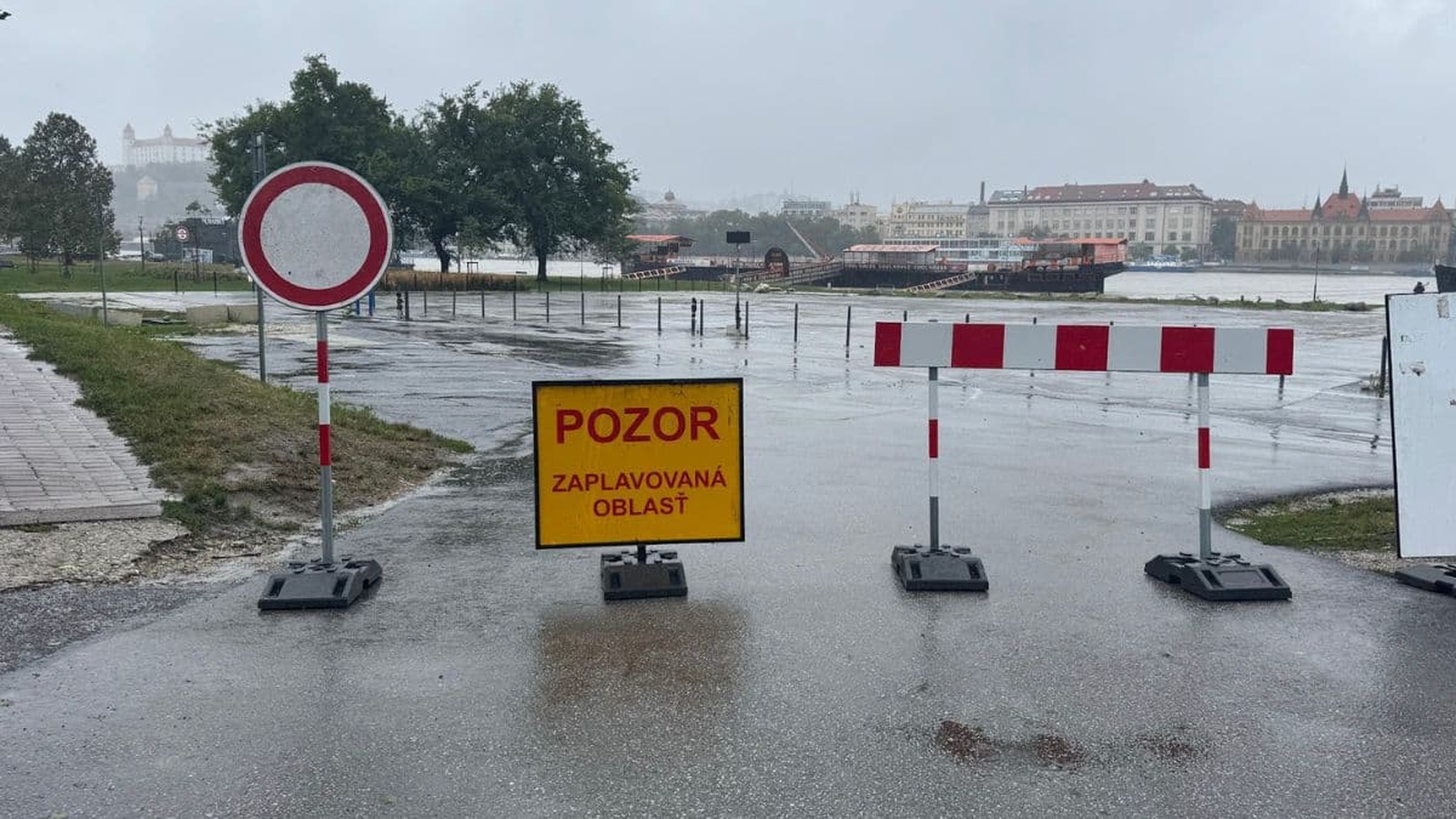 Záplavová oblasť označená dopravnou značkou a varovným žltým nápisom 'Pozor: zaplavovaná oblasť,' s pohľadom na zaplavenú cestu a Bratislavský hrad v pozadí.