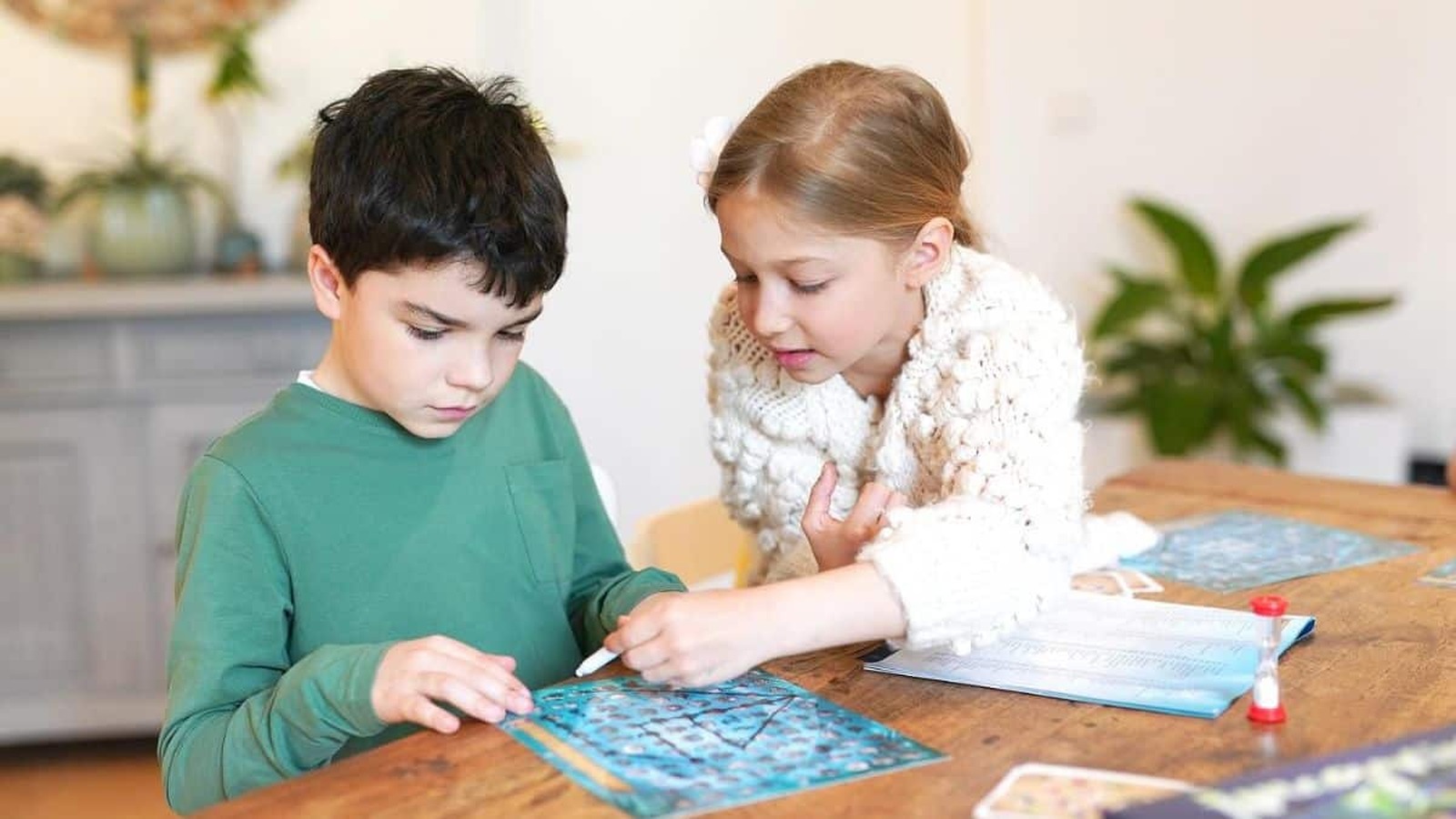 Dve deti sedia pri stole, kde sa venujú spoločenskej hre. Chlapec v zelenej mikine sa sústredene pozerá na hraciu dosku, zatiaľ čo dievča v bielej pletenej bunde mu niečo ukazuje a radí.