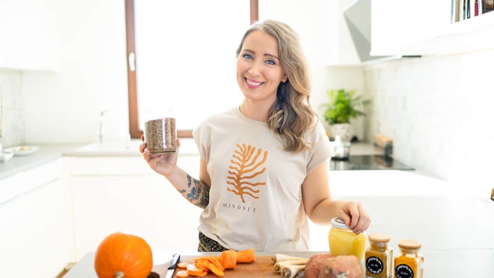 Mladá žena s úsmevom stojí v modernej kuchyni, drží pohár so semienkami, na stole má tekvicu, mrkvu a ďalšie čerstvé ingrediencie. Oblečená je v tričku s motívom "Mindset."