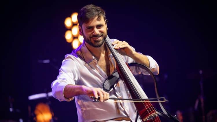 toznámy violončelista HAUSER, člen dua 2Cellos, hrá na elektrickom violončele počas koncertu, oblečený v bielej košeli, s úsmevom na tvári, v osvetlenej koncertnej sále.