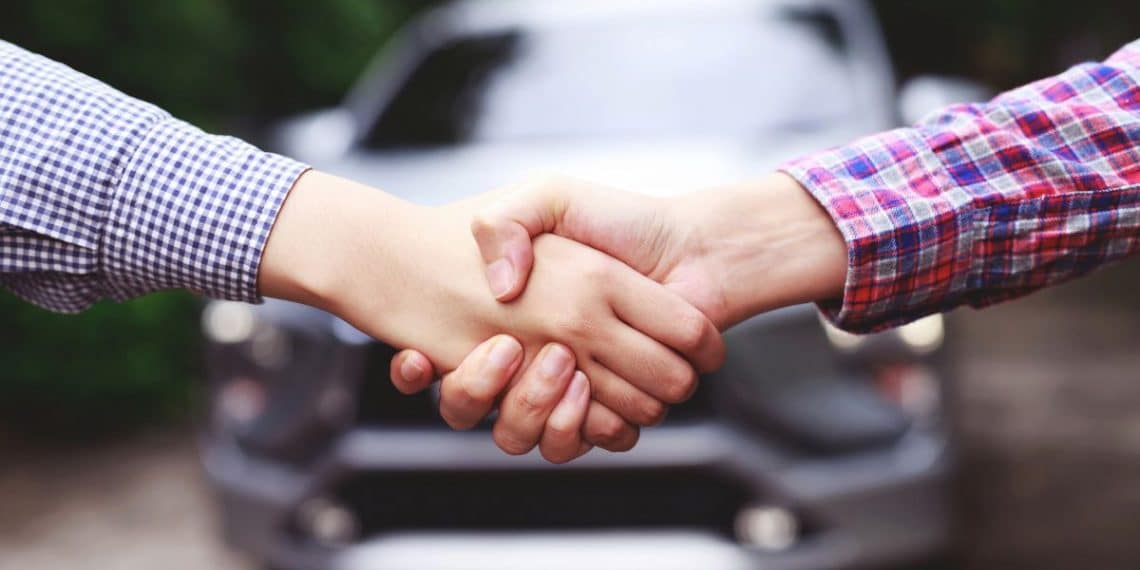 Podanie rúk medzi dvoma osobami, symbolizujúce dohodu pri predaji auta. V pozadí je viditeľný automobil.