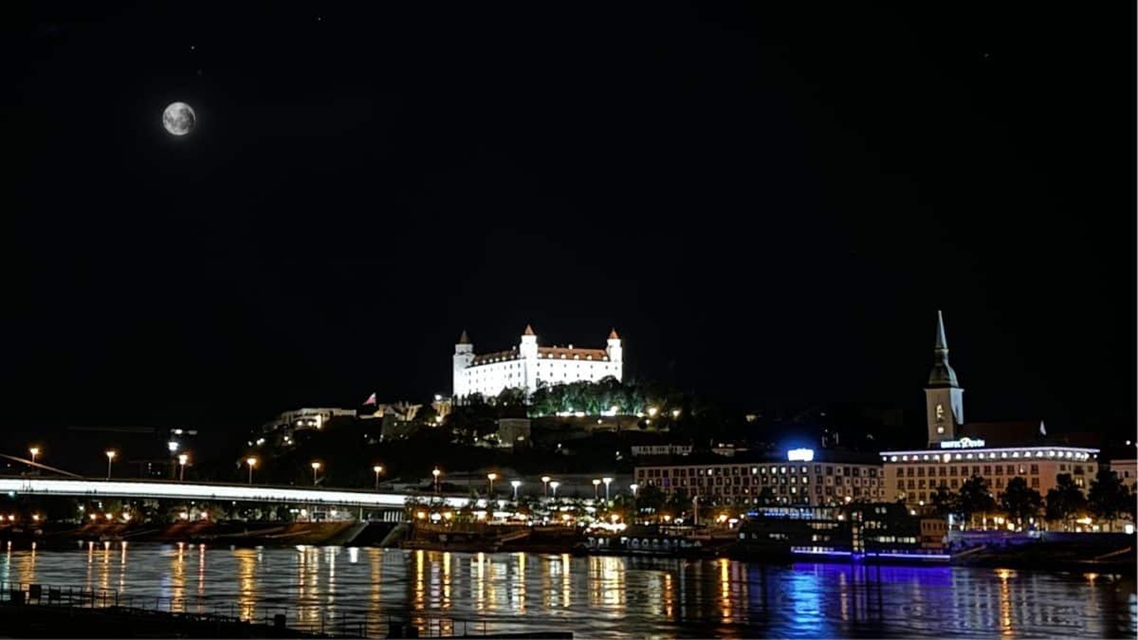 Nočný pohľad na osvetlený Bratislavský hrad a Most SNP s jasným splnom Mesiaca na oblohe.