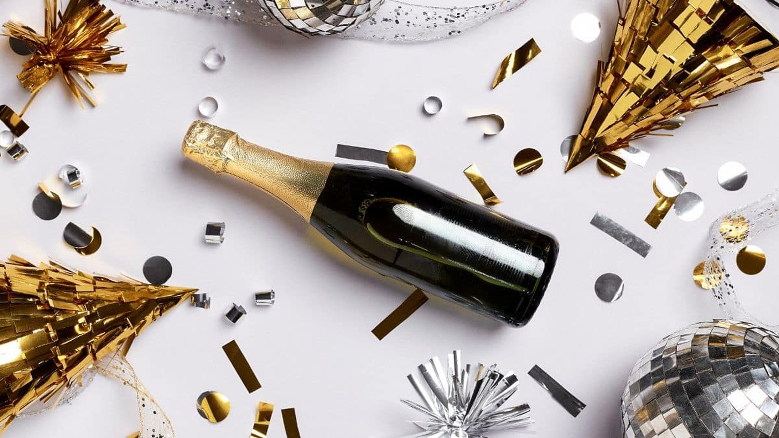 Fľaša šampanského obklopená zlatými a striebornými dekoráciami, konfety a party čiapkami na bielom pozadí.