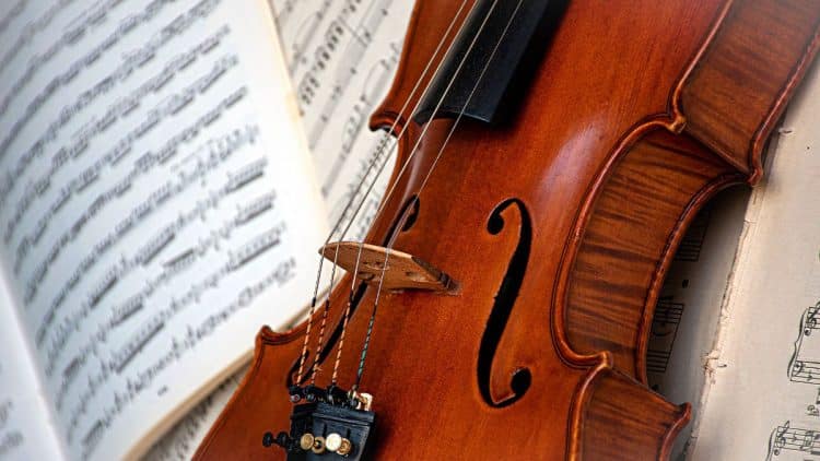 Detail violončela s partitúrou v pozadí, zobrazujúci jemné drevené textúry nástroja a struny, ktoré dodávajú obrazu hudobnú atmosféru.