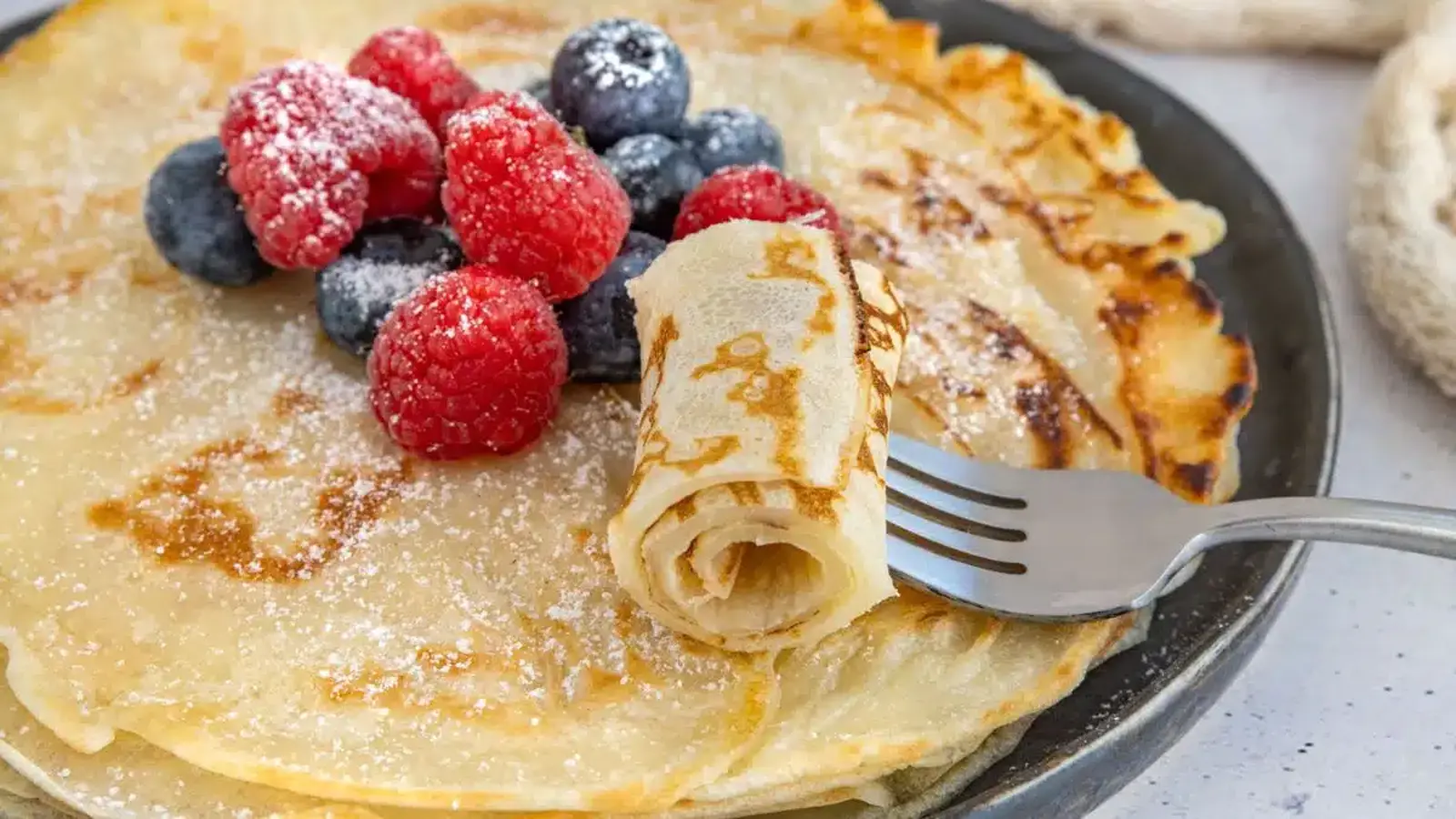 Detailný pohľad na zlatisté palacinky s čerstvými malinami a čučoriedkami, posypané práškovým cukrom, podávané na čiernom tanieri.