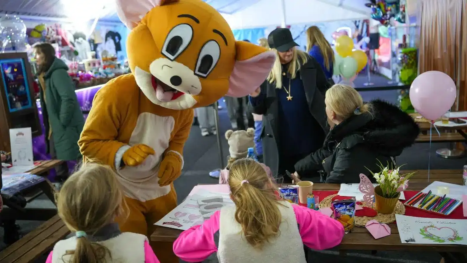 Deti sa zabávajú pri valentínskych tvorivých dielničkách v zábavnom parku, zatiaľ čo ich víta maskot v kostýme myši. Stoly sú plné pasteliek, omaľovánok a valentínskych dekorácií, v pozadí je veselo vyzdobený interiér s balónmi a svetielkami.