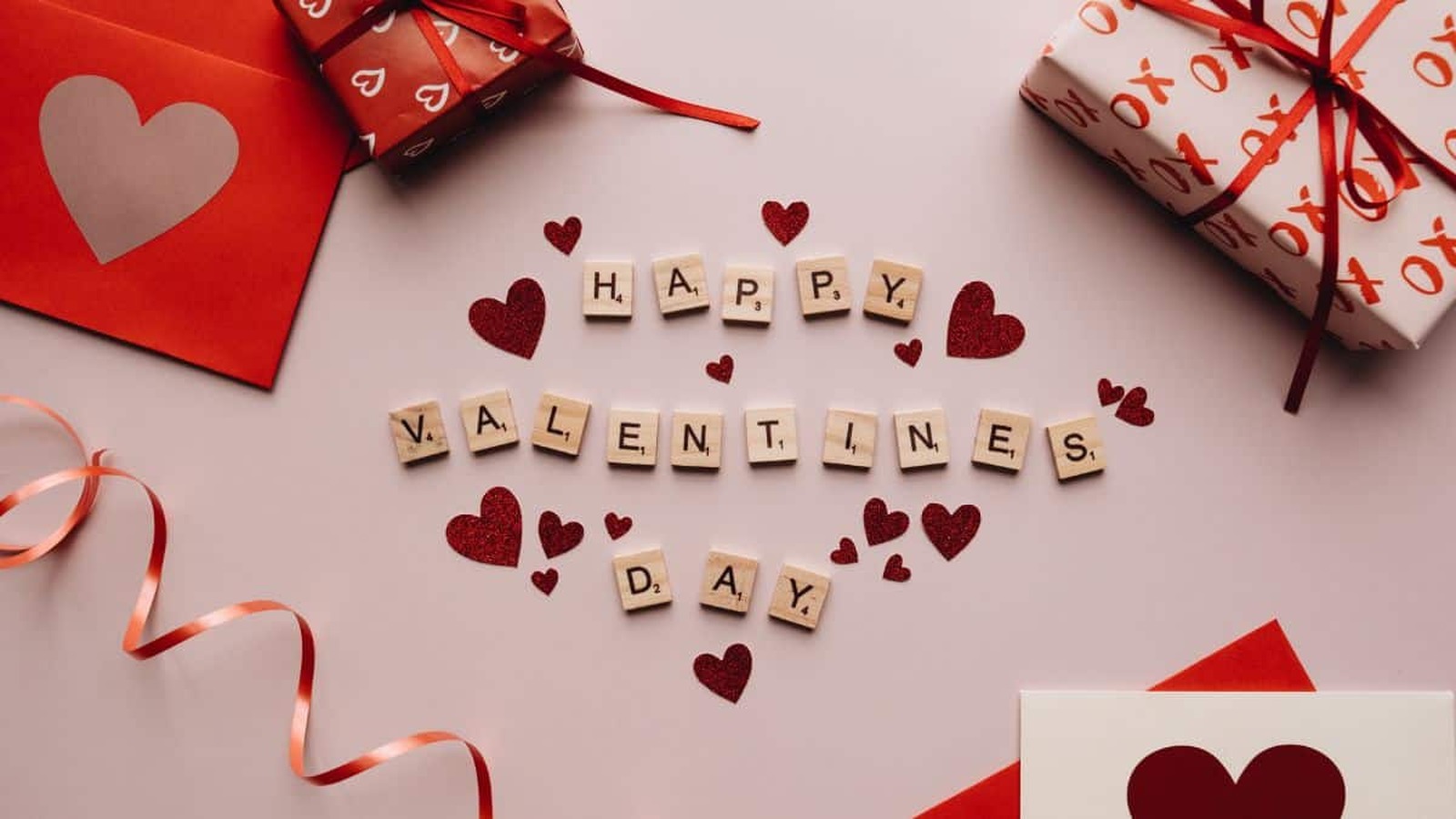 Valentínska dekorácia s nápisom „Happy Valentine's Day“ vytvoreným z drevených písmenkových kociek, obklopená červenými trblietavými srdiečkami, darčekmi a valentínskymi pohľadnicami.