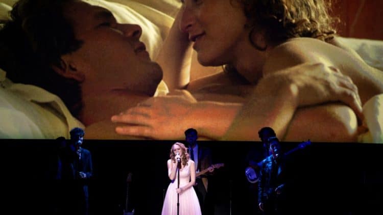 Speváčka a kapela na pódiu počas podujatia Dirty Dancing in Concert, na pozadí scéna z filmu Hriešny tanec s Patrickom Swayzem a Jennifer Grey.