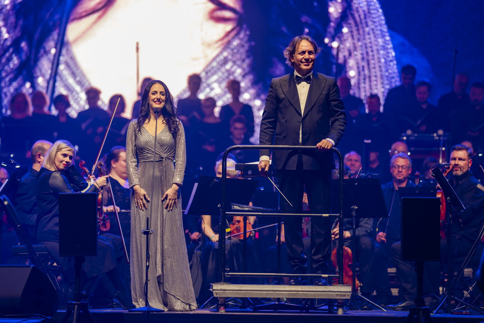 Dirigent a sólistka pred orchestrom na osvetlenom koncertnom pódiu